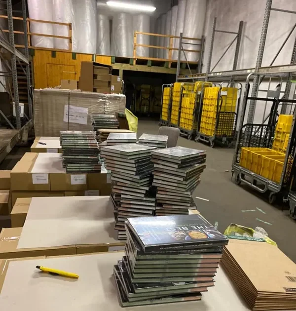 Packtisch mit Bücherstapeln in der Halle, dahinter gelbe Rollcontainer der Post, November 2019