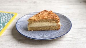 Bienenstich low carb: ein Stück mit sichtbarer Schichtung aus Mandelboden, Creme und Mandelkruste, im Hintergrund der angeschnittene Kuchen