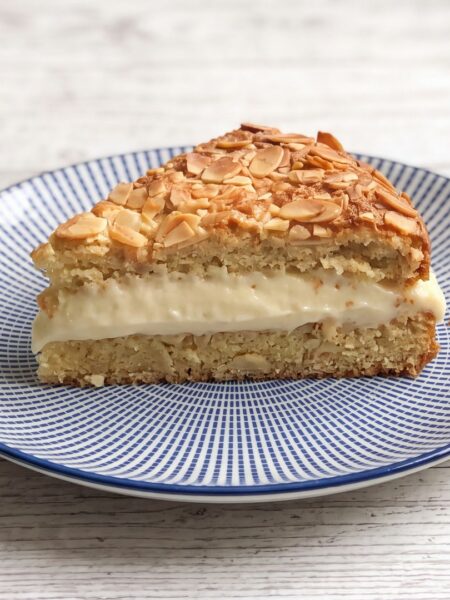 Bienenstich low carb: ein Stück mit sichtbarer Schichtung aus Mandelboden, Creme und Mandelkruste, im Hintergrund der angeschnittene Kuchen